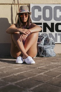 A stylish young woman sits casually on the ground, wearing sunglasses and a hat. Her backpack is by