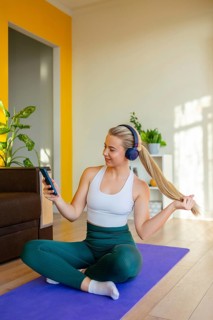 A woman in active wear sits on a yoga mat at home, wearing headphones and looking at her phone. She