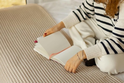 A person in a cozy, striped sweater sits cross-legged on a bed with a textured blanket, holding an