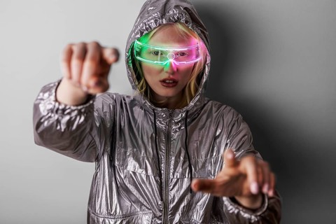 A woman wearing LED glasses and a metallic jacket poses against a neutral background. The bright
