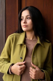 A woman stands against a wooden background, wearing a green jacket and brown top. Her gaze is