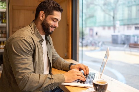 Man smiling and working on a laptop at a wooden table by a window in a cozy coffee shop. A cup of