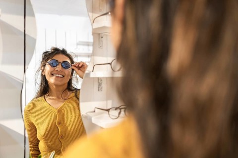 A smiling woman in a mustard sweater tries on stylish sunglasses while looking in a mirror at a