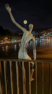 Night view of a sculptural figure on a river with a glowing tethered balloon in the background,