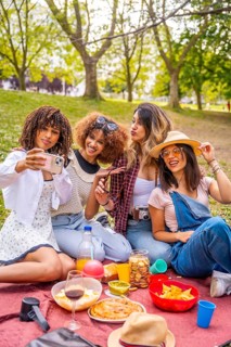 Four diverse female friends are enjoying a picnic in a park, taking a selfie with a smartphone