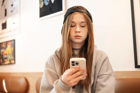 A teenage girl in a casual hoodie and cap sits indoors, focused on her smartphone. The background