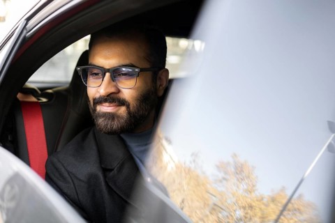 An Indian man dressed in a dark winter coat and glasses is seen smiling gently while driving his