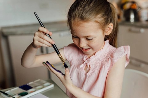 A joyful young girl in a pink top paints her hand with watercolor, showcasing creativity and fun.