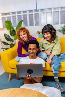 Vertical photo of three multiracial Lgtb people using laptop sitting together in a modern sofa