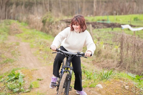 A young girl enjoys biking on a dirt trail, surrounded by greenery. She wears a cozy sweater and