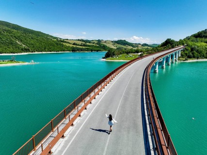 A young woman walks along a picturesque bridge overlooking a vibrant turquoise lake, surrounded by