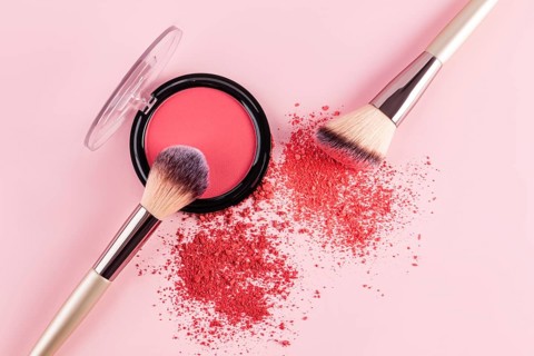 A vibrant pink compact blush and two makeup brushes are displayed on a soft pink background. The