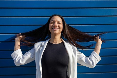A smiling Hispanic woman in a business outfit poses confidently against a blue backdrop, reflecting