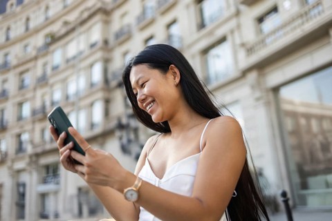 A young Chinese woman smiling while using her smartphone on a sunny summer day in the city. The