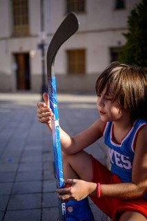 A young child sits on a paved street, holding a vibrant blue hockey stick. The scene captures a