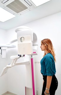 A young woman stands in a dental clinic, preparing for a panoramic dental X-ray. The advanced