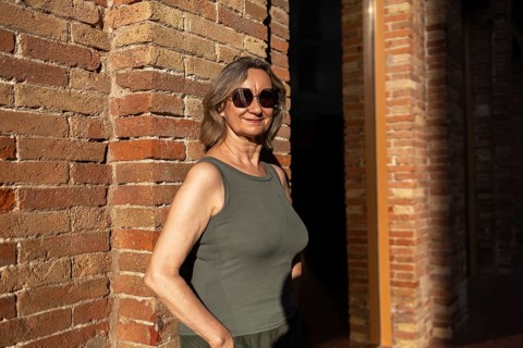 A joyful mature woman in casual attire stands against a brick wall, basking in the warm sunlight.