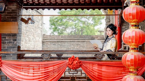 A woman in traditional Qing Dynasty attire, holding a fan, stands gracefully on a decorated balcony
