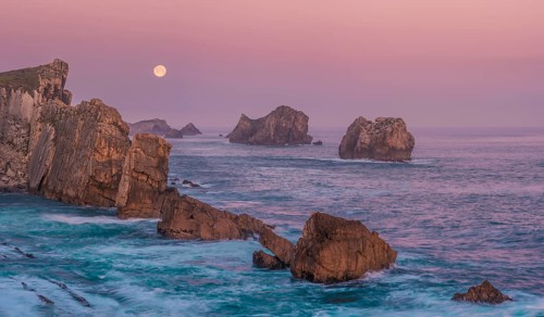 A serene view of rugged sea stacks and cliffs bathed in soft dawn light at Arnia Beach, Costa