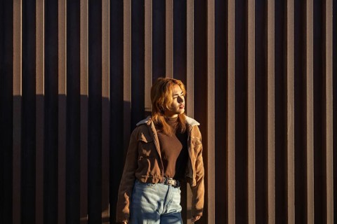 A Mixed-race woman stands against a vertical wooden wall, basking in warm sunlight She wears a