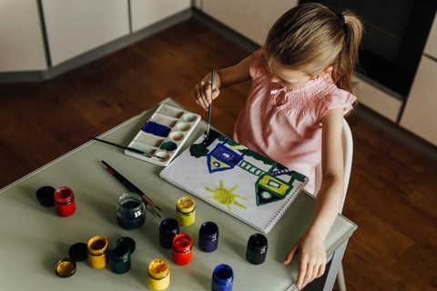 A child, dressed in a pink top, is intently painting a vibrant scene of houses and a sun using