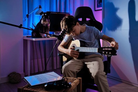 A young boy plays an acoustic guitar in a modern room with colorful lighting. A dog sits nearby on