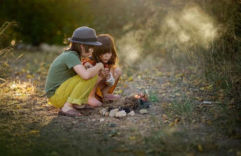 Two children sitting closely outdoors by a small campfire, surrounded by nature. They are focused