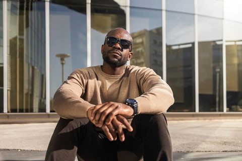 A confident cuban man wearing sunglasses and a casual beige sweater sits outdoors against a modern