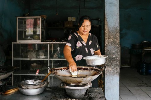 A woman prepares traditional food in a rustic kitchen of Bali, focusing intently as she cooks over
