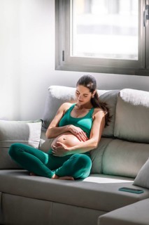 A pregnant woman peacefully sitting on a sofa in a sunlit room, dressed in a comfortable green yoga