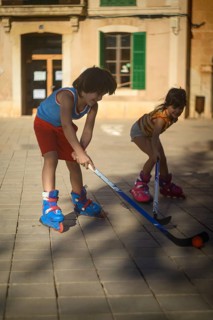 Two children play street hockey on a sunny day, wearing roller skates in an urban setting. The