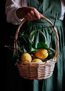 A close up of a wicker basket filled with fresh, ripe lemons, held by a person wearing a green