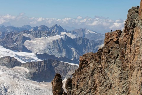 A hiker stands atop a rugged cliff in Zermatt, Switzerland, admiring the panoramic summer view of