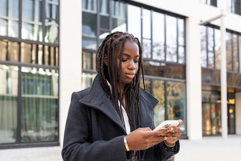A young African American woman with braids, dressed in a business coat, is using a smartphone while