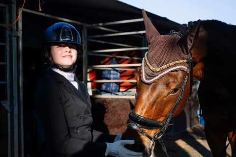 A rider in formal attire stands beside a brown horse, wearing a competition bridle and ear net The