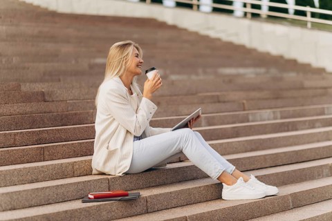 A businesswoman sits on outdoor steps with a tablet, enjoying a coffee. Dressed casually in white