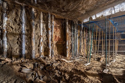 Underground construction site showing exposed dirt walls and temporary supports. The area is dimly