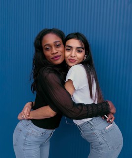 Two young women embrace in a joyful, friendly hug against a vibrant blue background. Their radiant