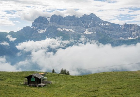 A serene view of a small cabin in a lush green meadow overlooking the Les Dents du Midi mountain