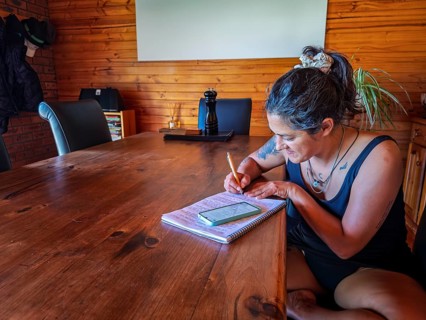 A woman is writing in a notebook at a large wooden table, surrounded by warm decor in Australia.
