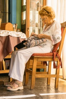 Elderly woman wearing glasses and white clothes is focused on sewing a colorful fabric while