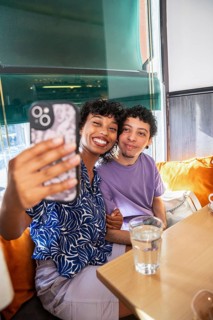 A joyful, multiethnic LGBTQ+ couple sits in a cozy cafe, smiling as they take a selfie. Sunlight