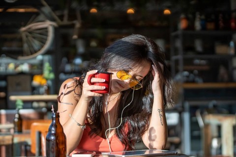 Woman in a cafe, savoring coffee with yellow sunglasses and earphones, enjoying music on a sunny