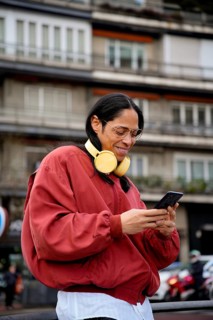 A man with long hair and large glasses wearing a red jacket and yellow headphones is outside