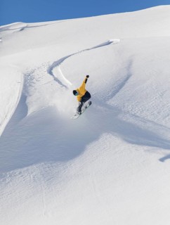A snowboarder in a yellow jacket executes a jump on a snowy mountain slope. The clear blue sky and