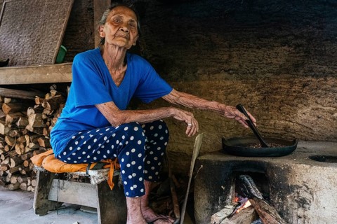 An elderly woman in a rustic kitchen prepares balinese coffee on a traditional wood fired stove.