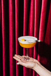 A peach cocktail served in an elegant coupe glass, delicately held against a backdrop of rich red