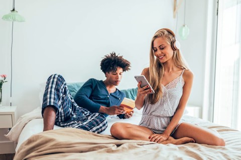 A couple enjoys a relaxed morning on a cozy bed, one listens to music on headphones, the other