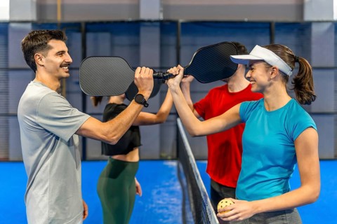 Four friends on a blue pickleball court paddle to paddle over the net, smiling and high fiving