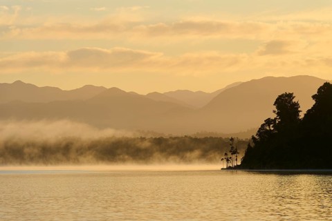 Mist drifts over a calm lake with mountains in the background at sunrise, Lake Mahinapua, Ruatapu,